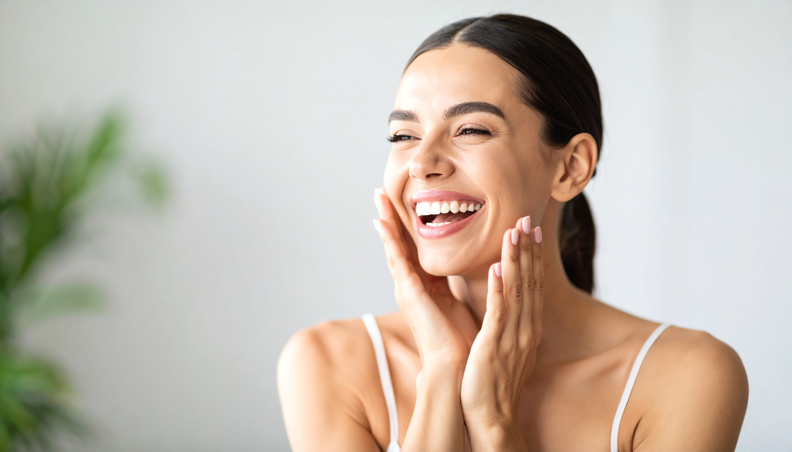 A young woman with dark hair pulled back smiles brightly, touching her cheeks with both hands. She is wearing a white tank top and appears happy and relaxed, enjoying the results of her recent facial, with a blurred green plant in the background.