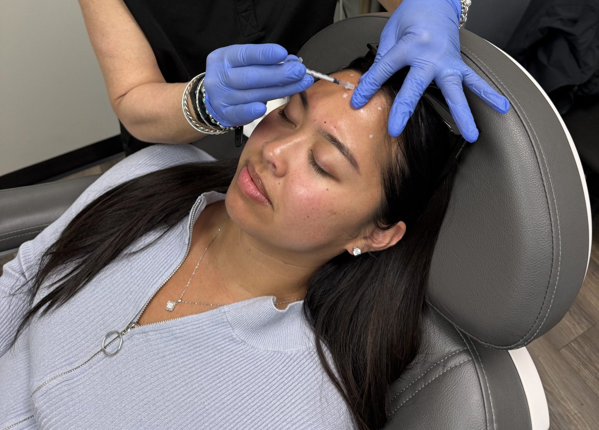 A woman lies on a chair with her eyes closed while a medical professional in blue gloves injects Botox into her forehead at dermani MEDSPA®.